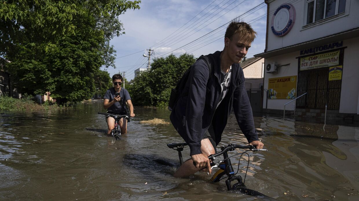 Местные жители пытаются проехать на велосипедах по затопленной дороге после прорыва плотины Каховской ГЭС в Херсоне, Украина, 6 июня 2023 года.