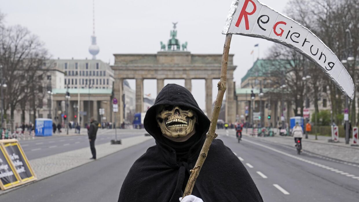 Мужчина, переодетый в смерть, протестует перед Бранденбургскими воротами с косой, на которой надписано Правительство, во время демонстрации фермеров в Берлине