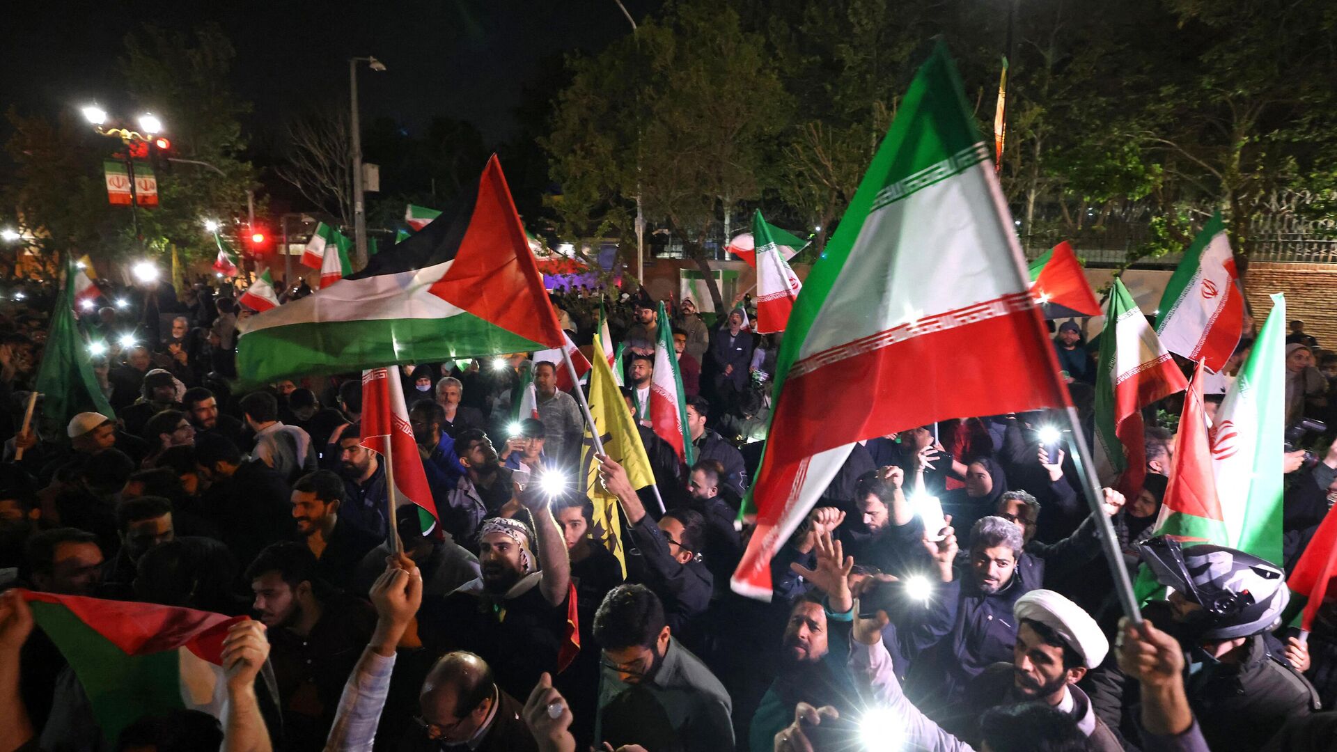 Демонстранты размахивают флагом Ирана и палестинскими флагами перед посольством Великобритании в Тегеране 14 апреля 2024 года после того, как Иран атаковал Израиль беспилотниками и ракетами - ИноСМИ, 1920, 15.04.2024