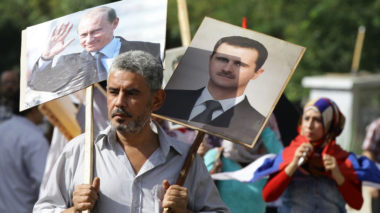 Сириец держит плакаты с изображениями Владимира Путина и Башара Асада на акции в поддержку российских военных действий в Сирии, Дамаск
