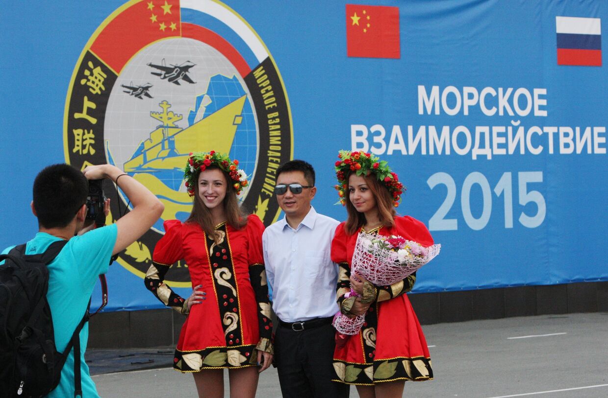 Участник китайской делегации фотографируется с русскими девушками на фоне баннера совместных учений Морское взаимодействие - 2015