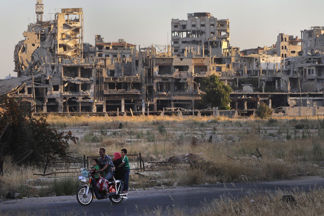 Люди едут на мотоцикле мимо поврежденных зданий в городе Хомс, Сирия
