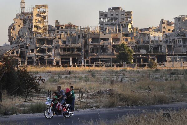 Люди едут на мотоцикле мимо поврежденных зданий в городе Хомс, Сирия