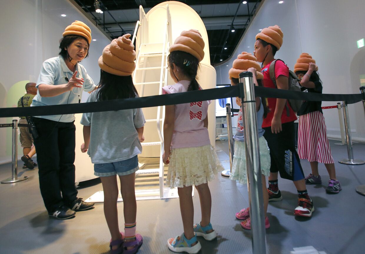 Экскурсовод в Национальном музее развития науки и инноваций в Токио рассказывает детям в шапочках в виде какашек о работе канализации
