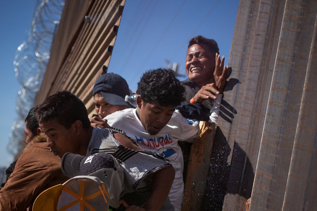 Мигранты пробираются через забор на границе, возвращаясь в Мексику после применения сотрудниками полиции слезоточивого газа в Тихуане