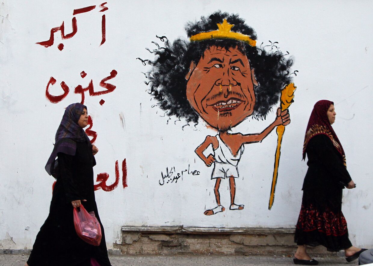 Женщины идут мимо граффити, изображающее Муаммара Каддафи