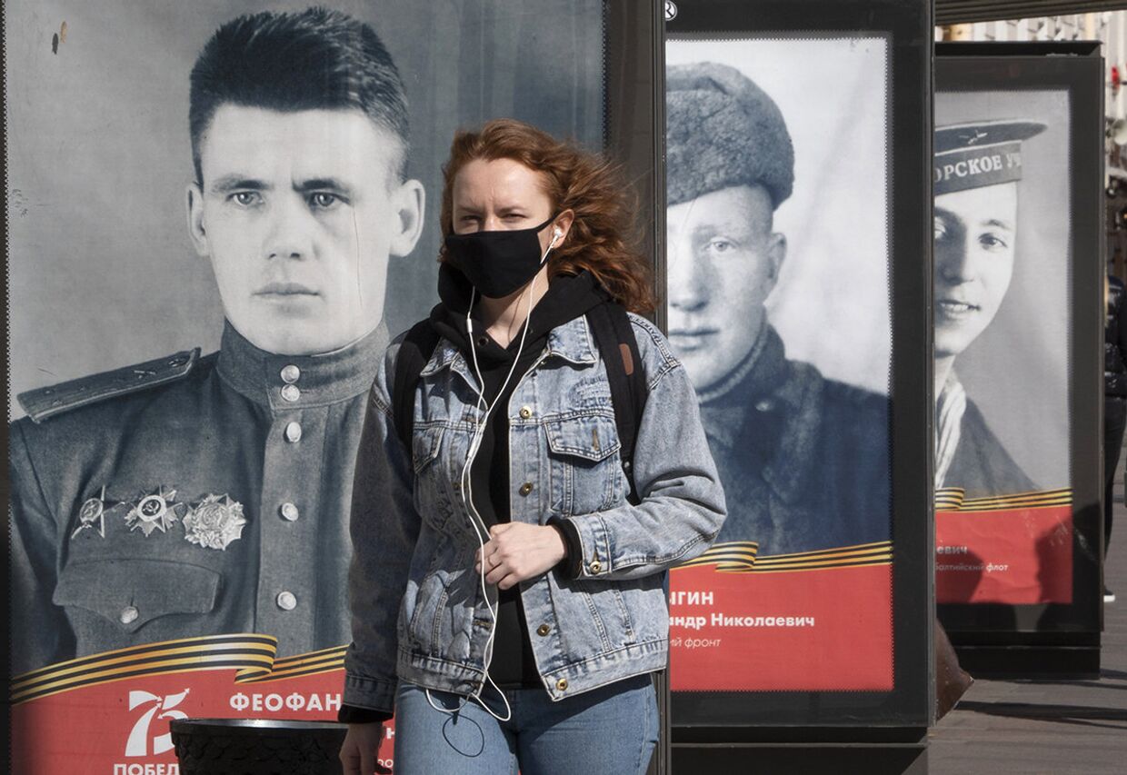 Женщина в маске идет мимо плакатов с портретами солдат Великой Отечественной войны